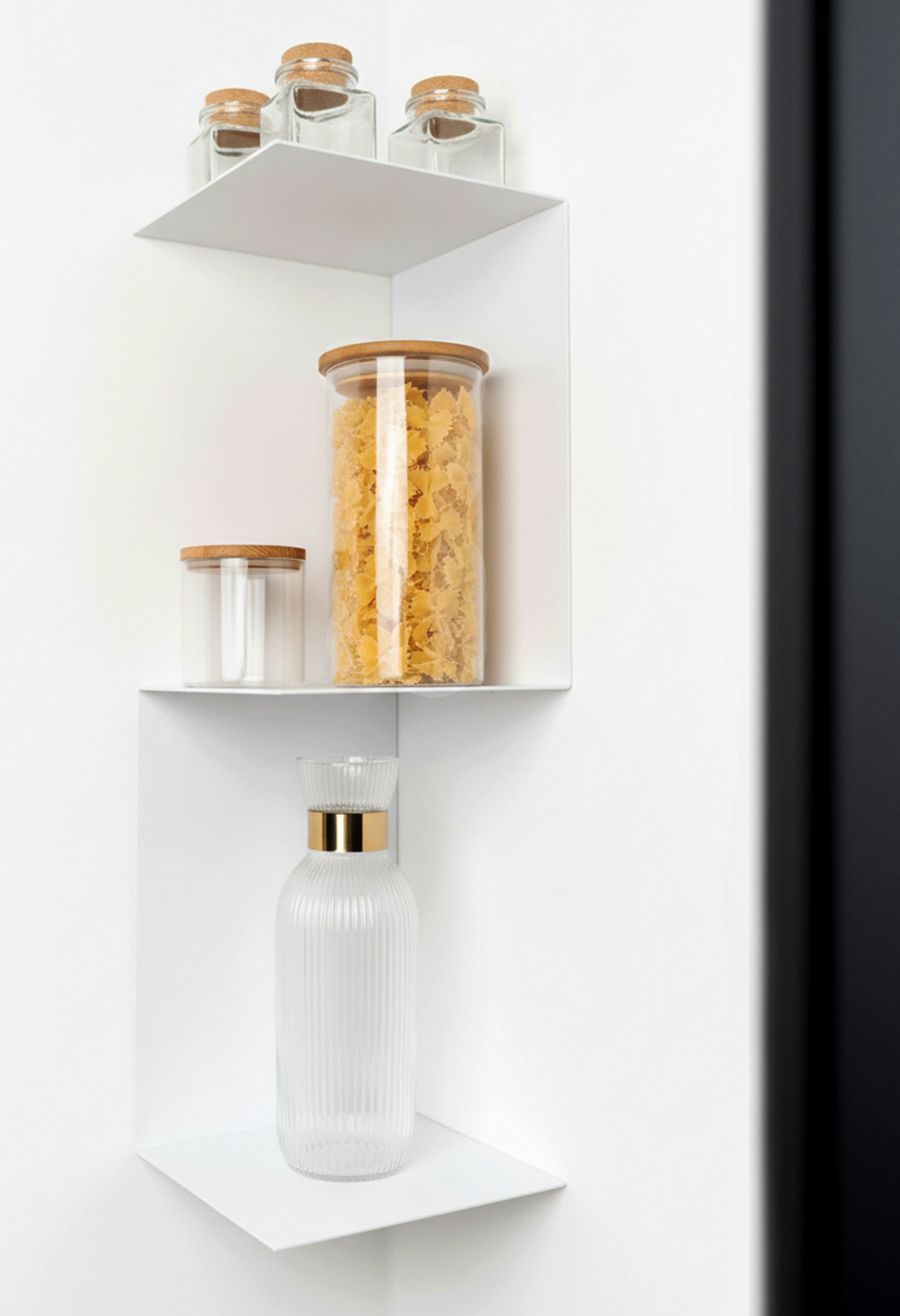 Weisses, minimalistisches Eckwandregal aus Metall, das in einer Ecke hängt und mit Glasgefäßen dekoriert ist. Die Gefäße enthalten Nudeln und Gewürze, eines der Gläser hat einen Holzdeckel. Das Regal bietet eine stilvolle und platzsparende Lösung für die Raumnutzung in der Küche
