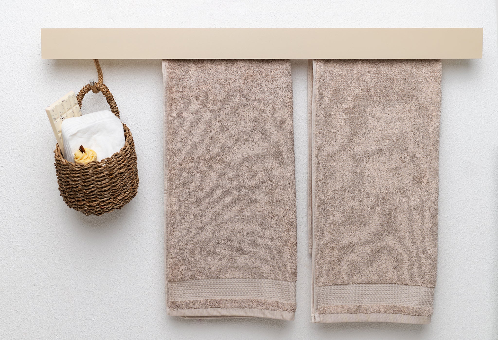 Beiger langer Metall Handtuchhalter mit schmaler Blende hinter welche zwei beige Handtücher hängen. Links hängt zudem ein Körbchen mit Kosmetikutensilien an einem beigen S Haken hinter der Blende hervor.
