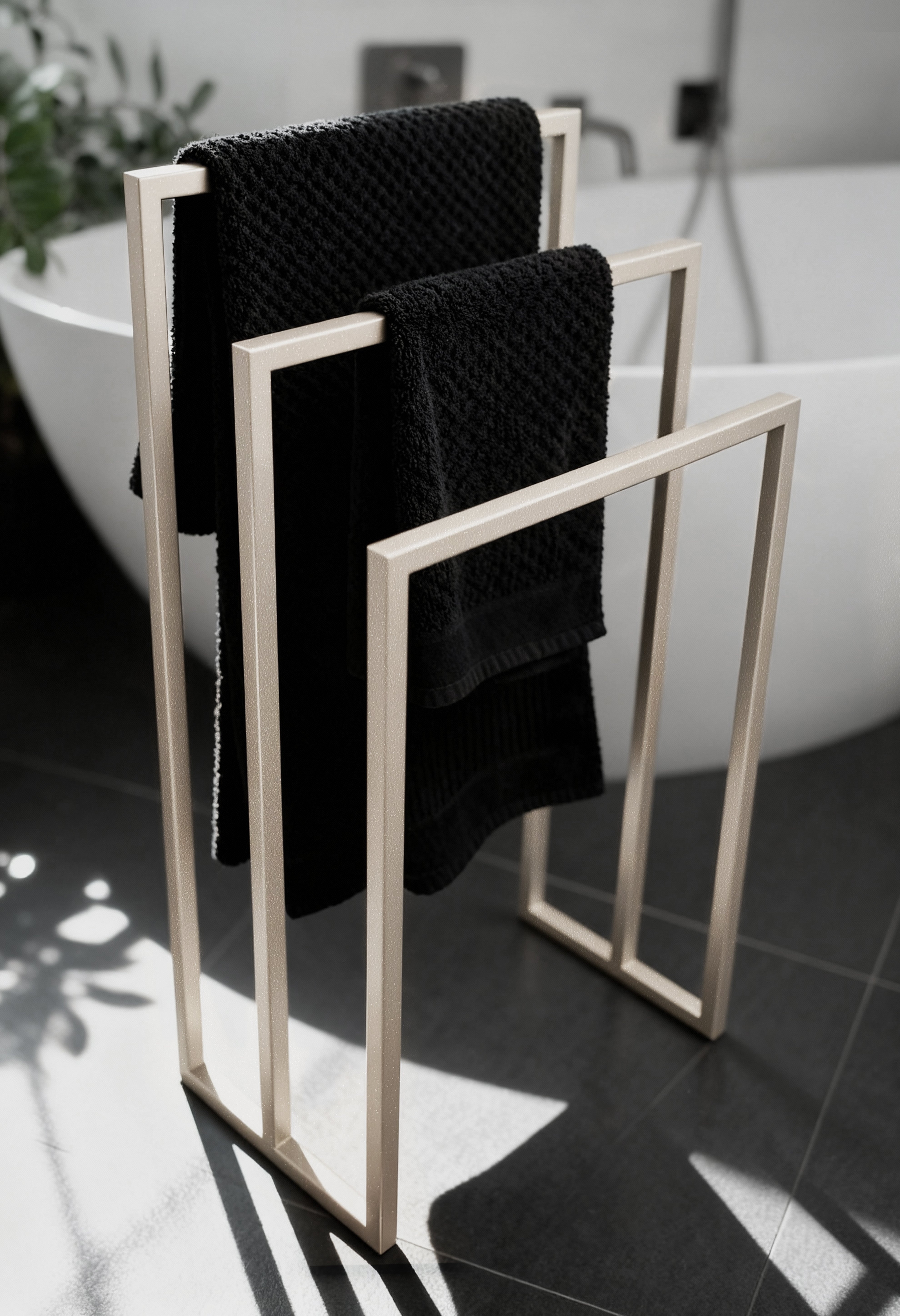 Freistehender Handtuchhalter aus Metall in champagnerfarben leicht glitzernd mit drei unterschiedlich hohen Stangen für Handtücher verschiedener Größen. Der Handtuchhalter steht vor einer modernen großen Badewanne in einem eleganten Badezimmer mit dunklen Fliesen