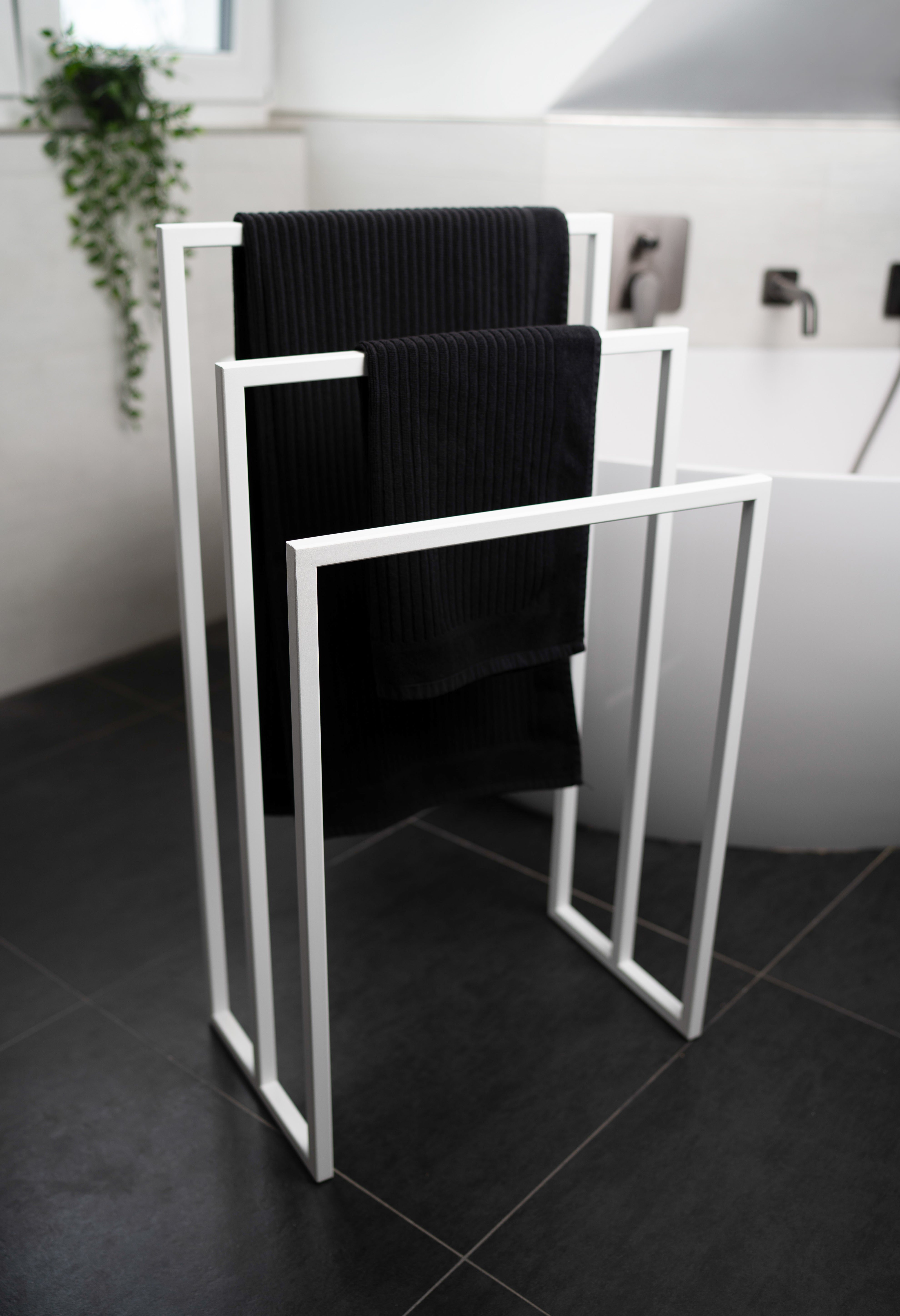 Freistehender Handtuchhalter aus weissem Metall mit drei unterschiedlich hohen Stangen für Handtücher verschiedener Größen. Der Handtuchhalter steht vor einer modernen großen Badewanne in einem eleganten Badezimmer mit dunklen Fliesen