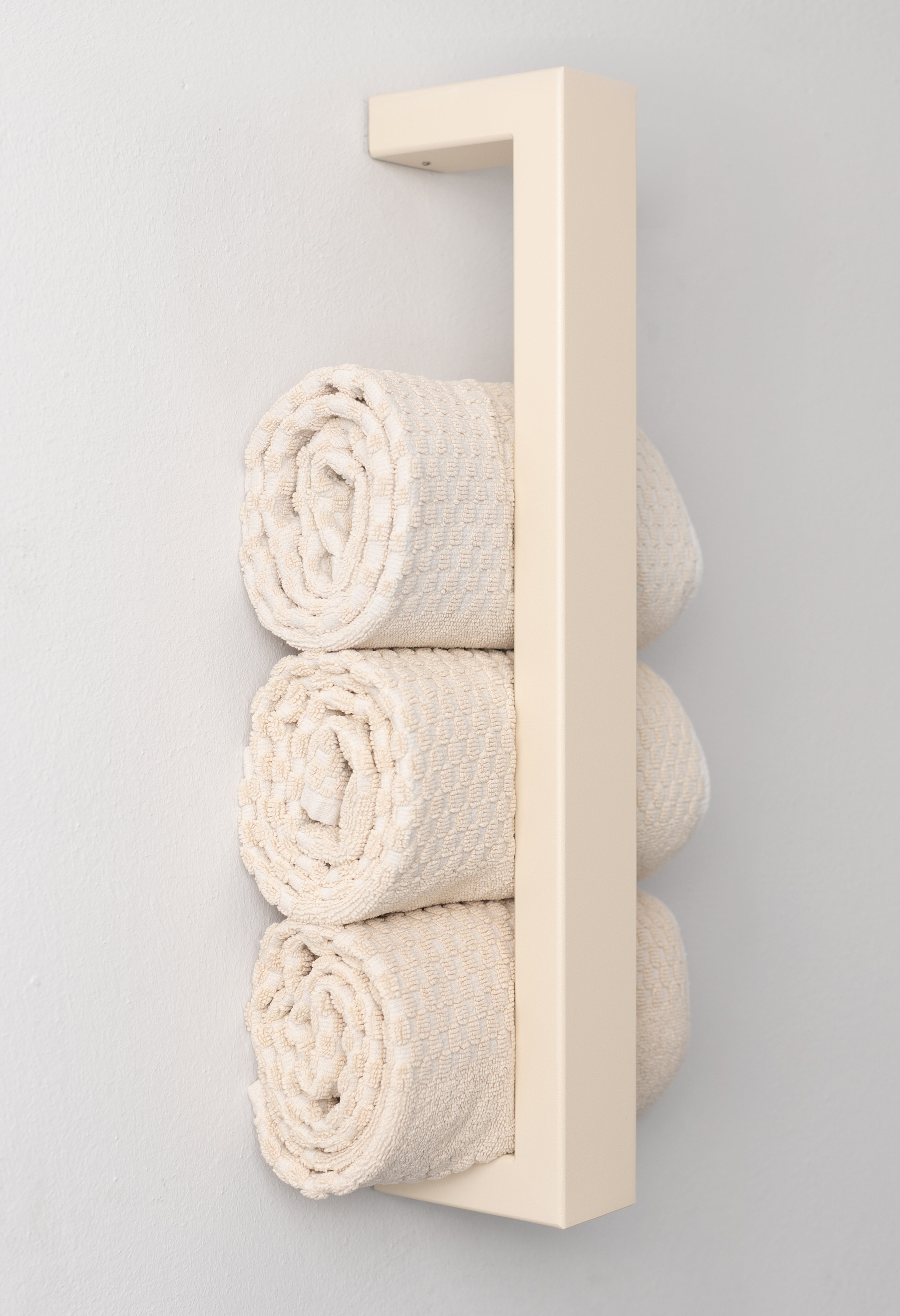 Beiger, moderner Handtuchhalter aus pulverbeschichtetem Metall, der an einer Wand montiert ist. Der Halter bietet Platz für mehrere gerollte Handtücher und fügt sich dekorativ in das Badezimmer ein. Eine stilvolle, platzsparende Lösung zur Aufbewahrung von Handtüchern.