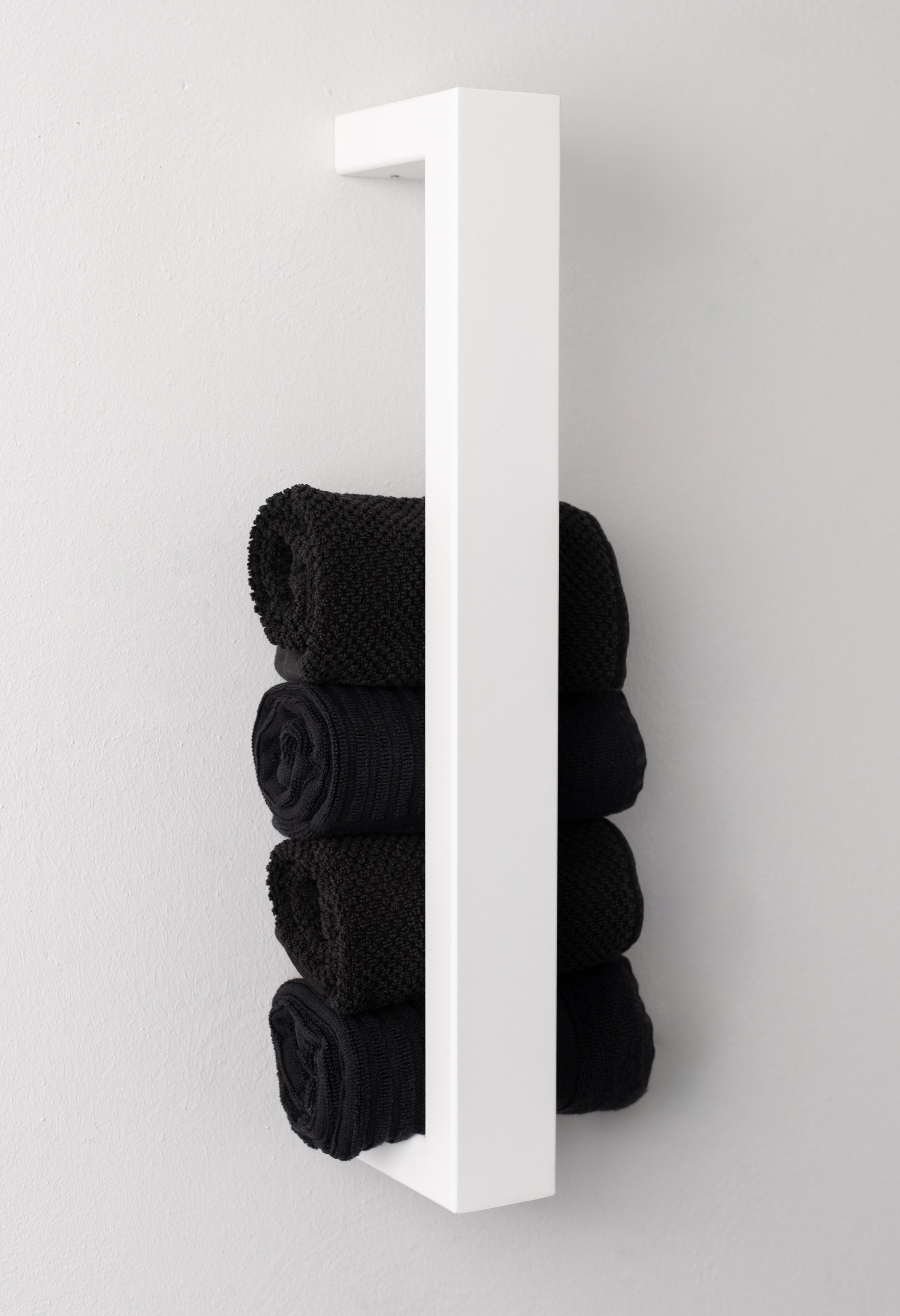 Weisser, moderner Handtuchhalter aus pulverbeschichtetem Metall, der an einer Wand montiert ist. Der Halter bietet Platz für mehrere gerollte Handtücher und fügt sich dekorativ in das Badezimmer ein. Eine stilvolle, platzsparende Lösung zur Aufbewahrung von Handtüchern.