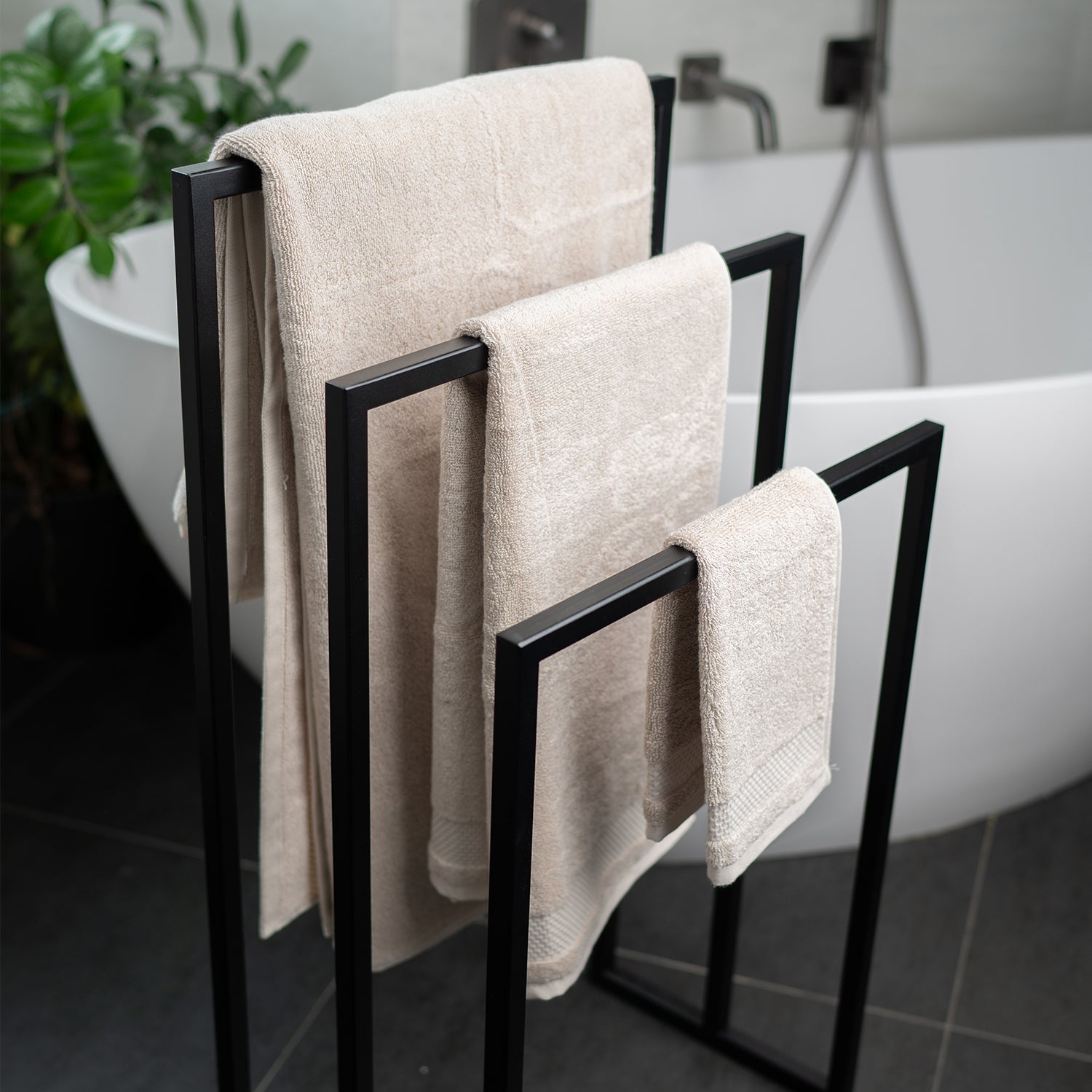 Stehender Handtuchhalter schwarz pulverbeschichtet aus Metall, vor weißer moderner Badewanne. Der rechteckige Handtuchhalter hat 3 verschiedene Höhen und es hängen passend dazu drei verschiedene Handtuchgrößen auf.