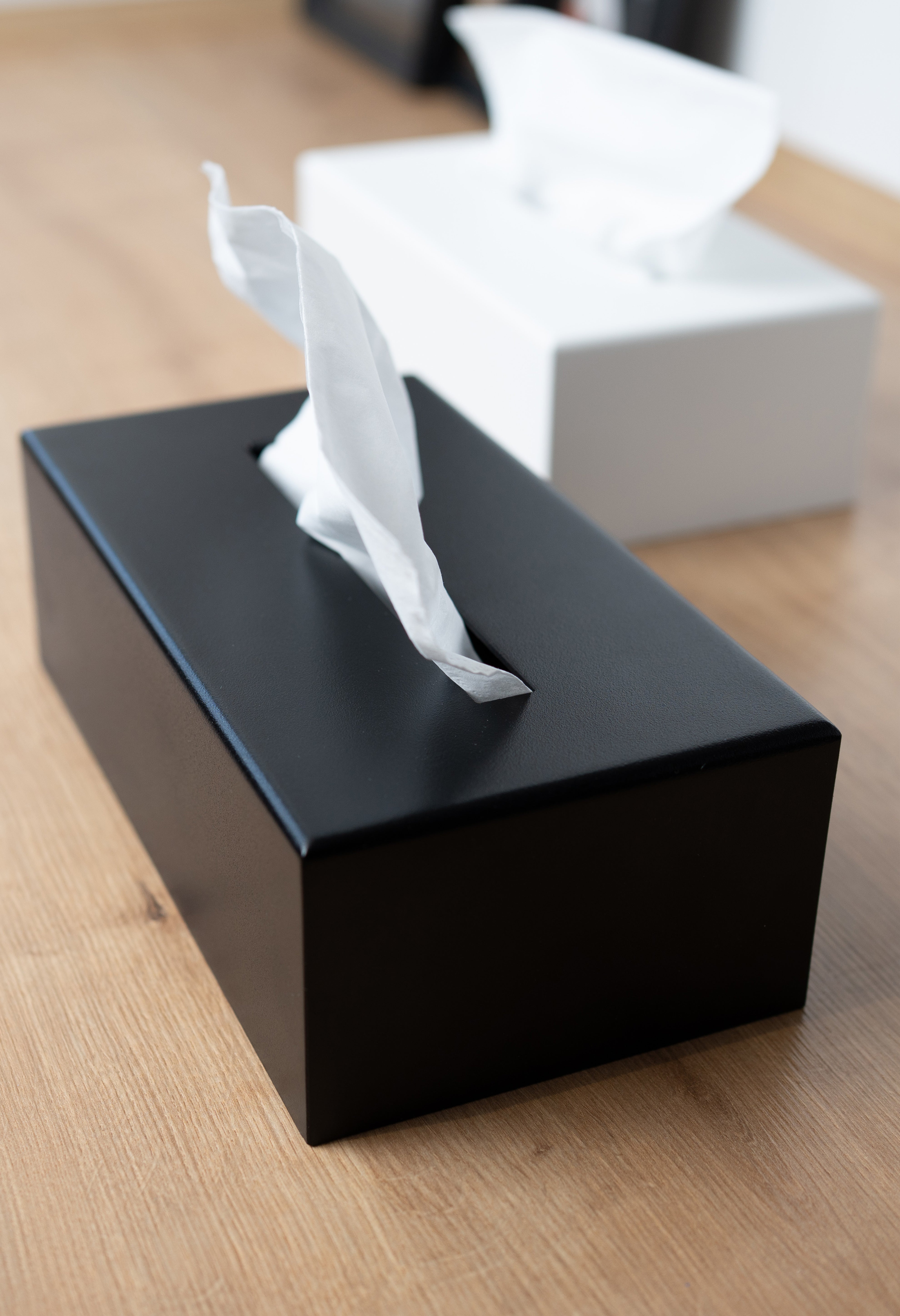 Kosmetiktuchbox in schwarz und weiß, die schwarze Box im Fokus und im Hintergrund sieht man die weiße Box beide stehen auf einer hellen Holzfläche.