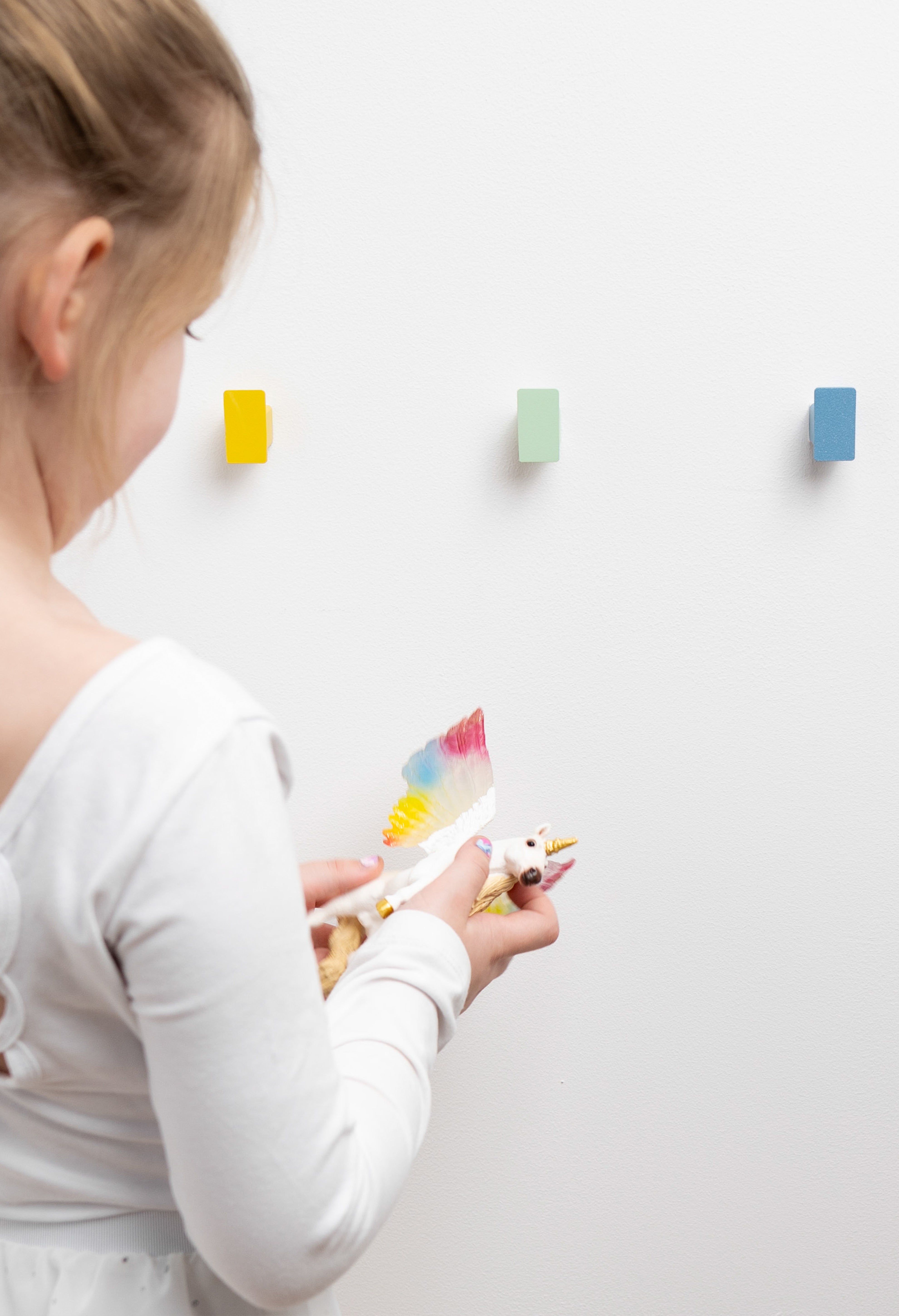 Farbenfrohe pulverbeschichtete Design Kindergarderobe aus Metall mit sechs bunten Haken in Regenbogenfarben im Close-Up Ein süßes 5jähriges Mädchen steht vor der Garderobe und hält ein kleines Spielzeug Einhorn, von welchem die Flügelfarben exakt zu den Garderobenhaken von CAPRI passen.