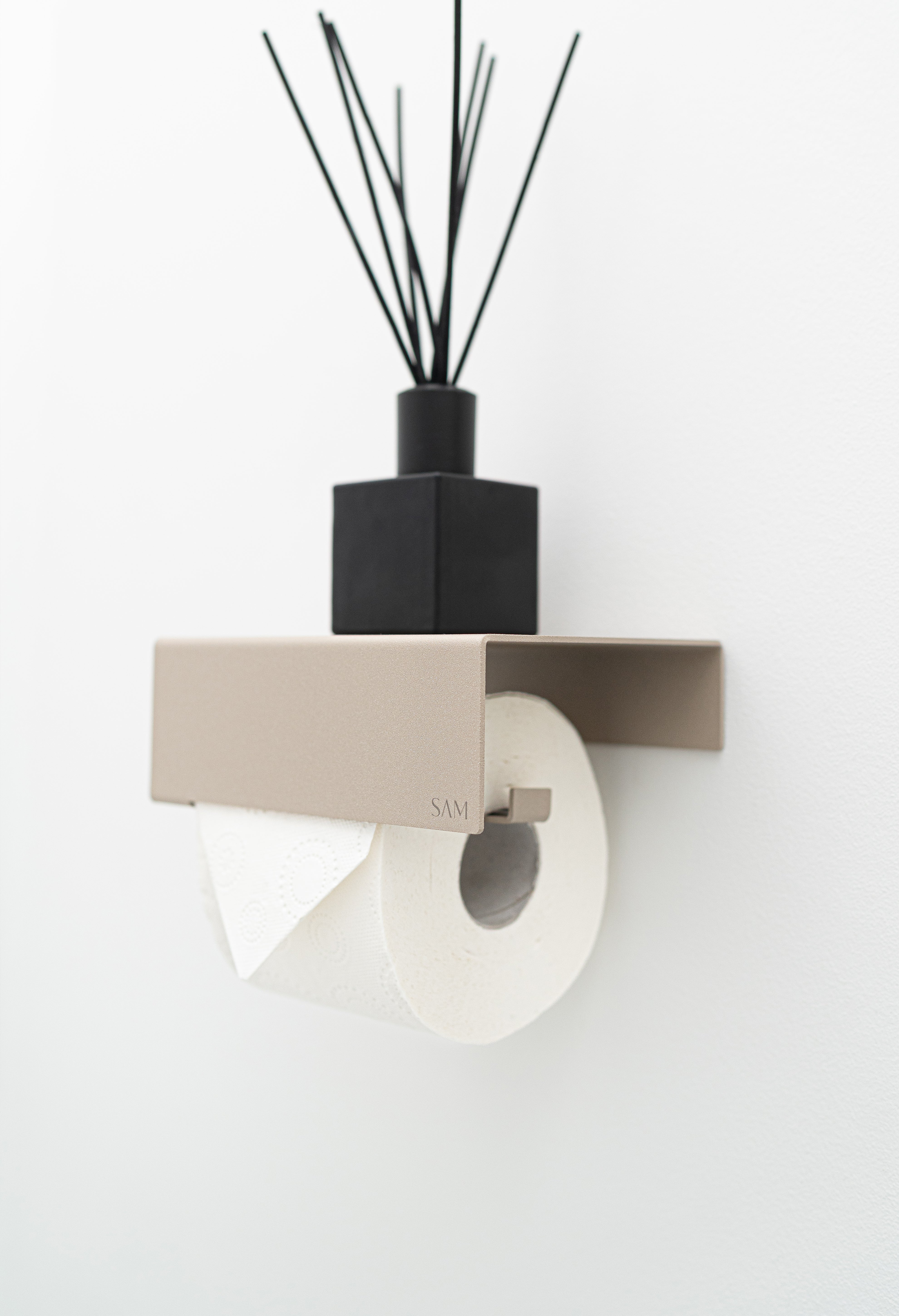 Toilettenpapierhalter LILLE zum Kleben ohne Bohren in Farbe Champagner in schräger Ansicht mit einer toilettenpapierrolle bestückt und einem Raumduft auf der Ablage.
