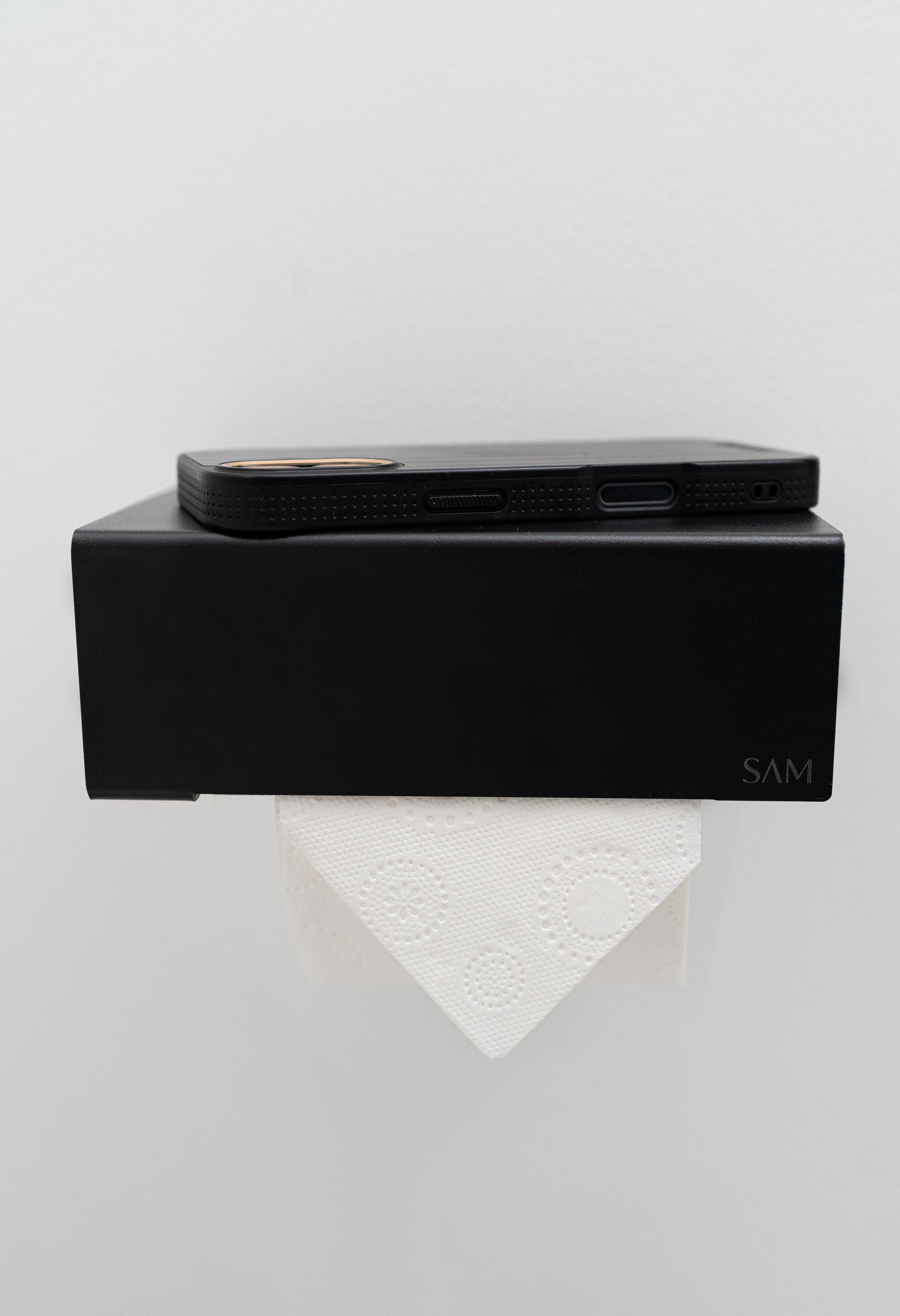 Toilettenpapierhalter LILLE zum Kleben ohne Bohren in Frontalansicht in Farbe schwarz mit Iphone, welches auf der Ablage liegt.