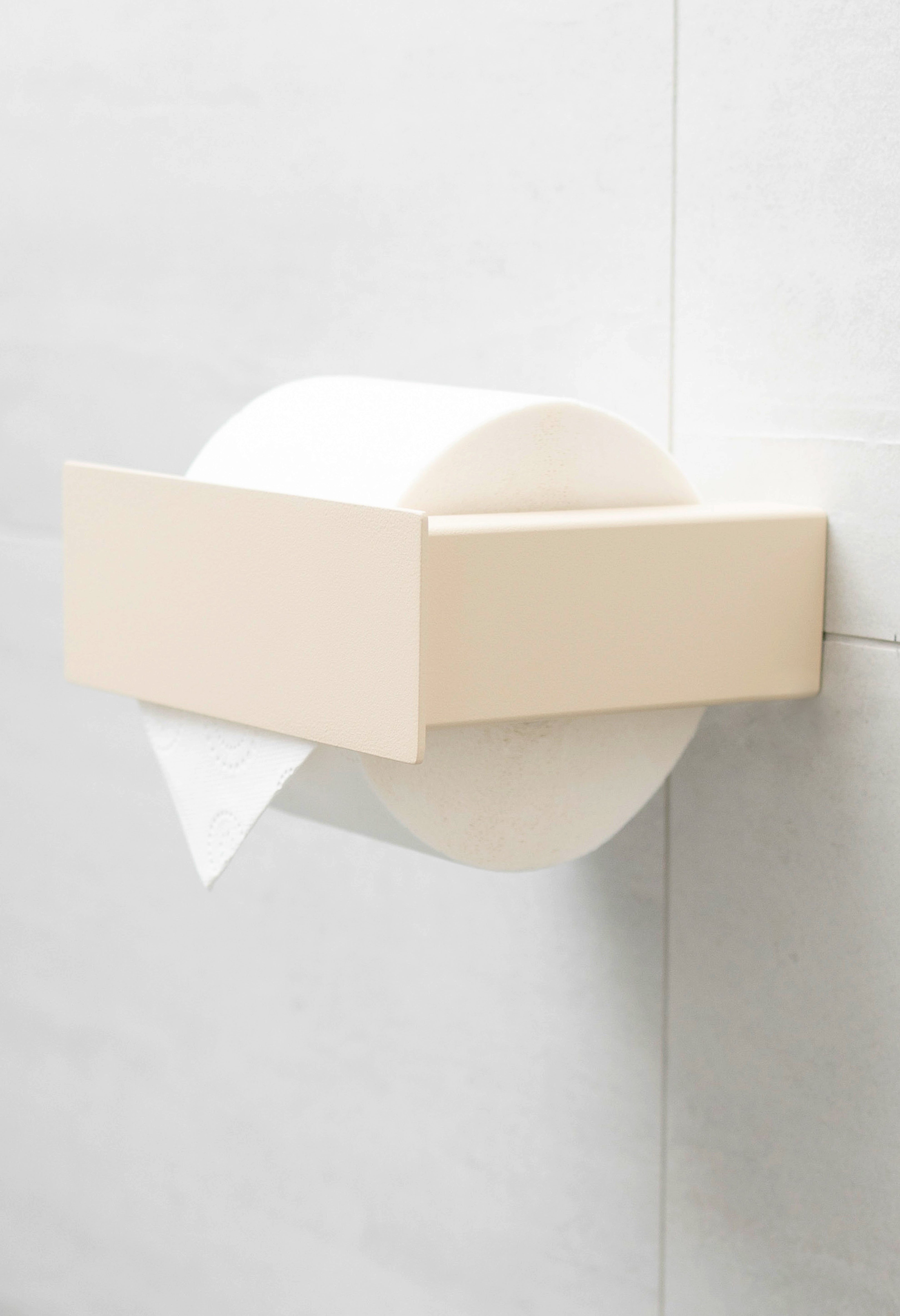 Beiger pulverbeschichteter Toilettenpapierhalter aus Metall mit Blende an weißen Fliesen.