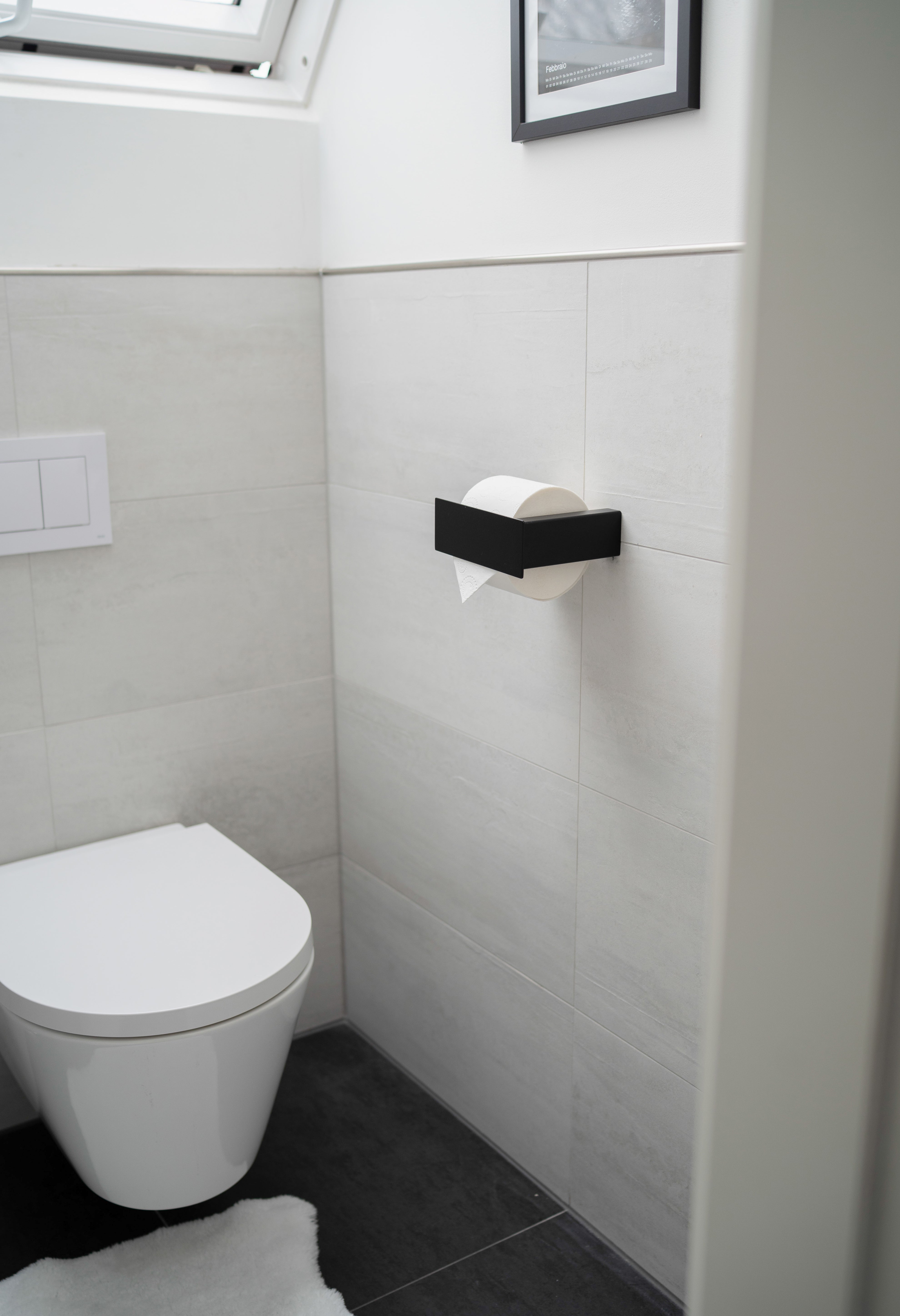 Schwarzer pulverbeschichteter Toilettenpapierhalter aus Metall mit Blende in kleinem hellem Badezimmer neben Toilette aufgehängt.