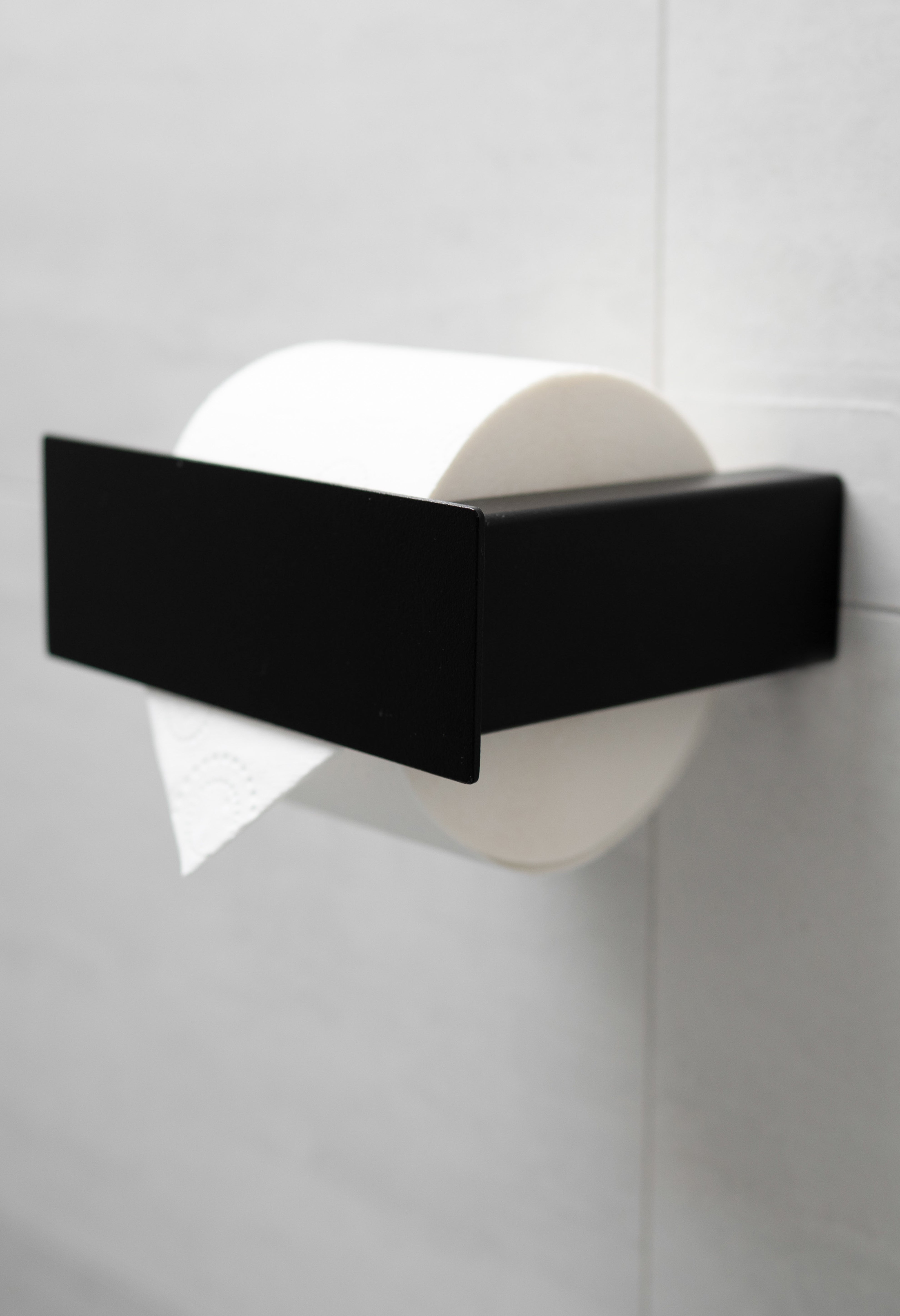 Schwarzer pulverbeschichteter Toilettenpapierhalter aus Metall mit Blende an weißen Fliesen.