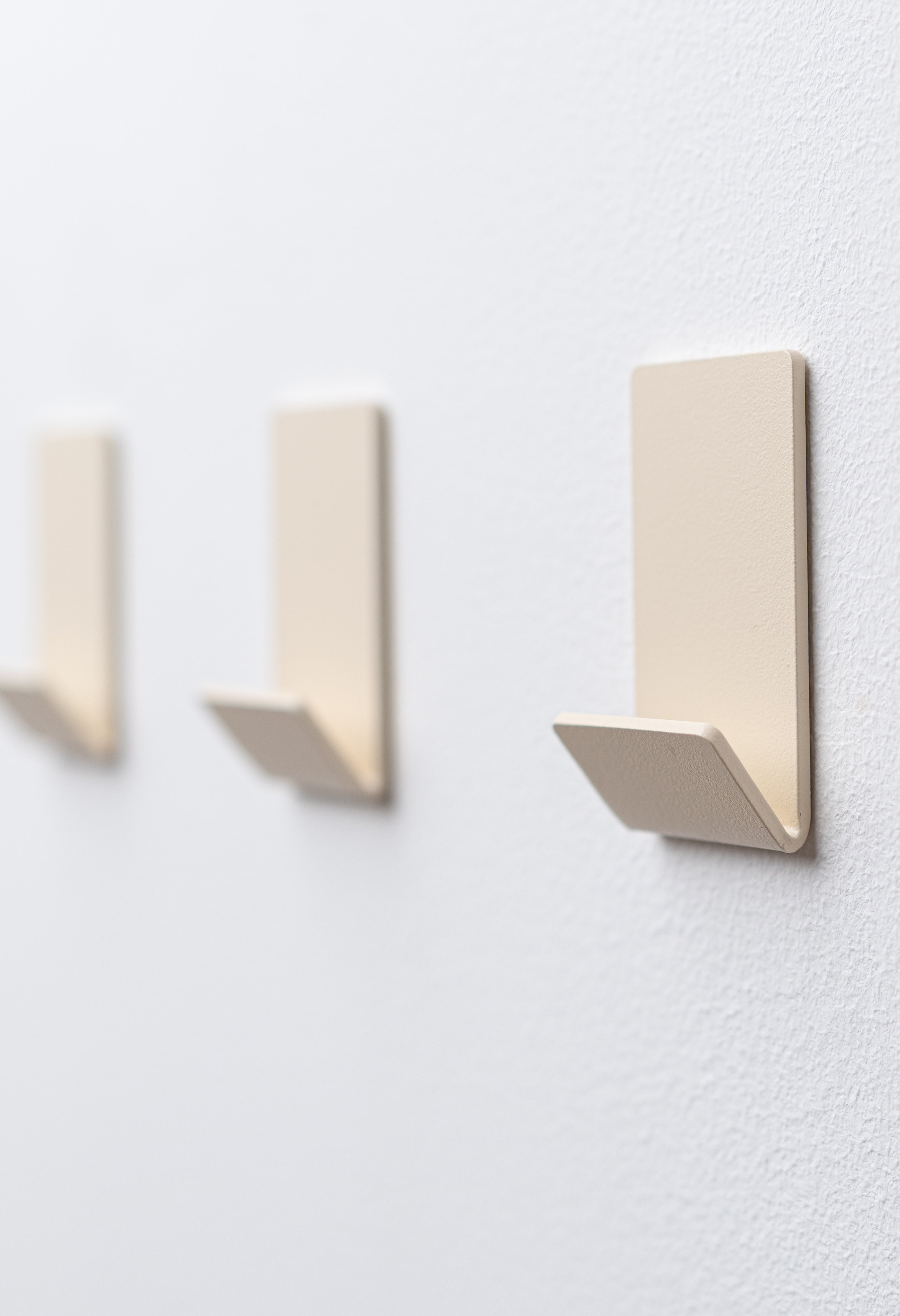 Wandhaken zum Kleben Ohne Bohren in Beige pulverbeschichtet als dreier Set an weißer Wand.