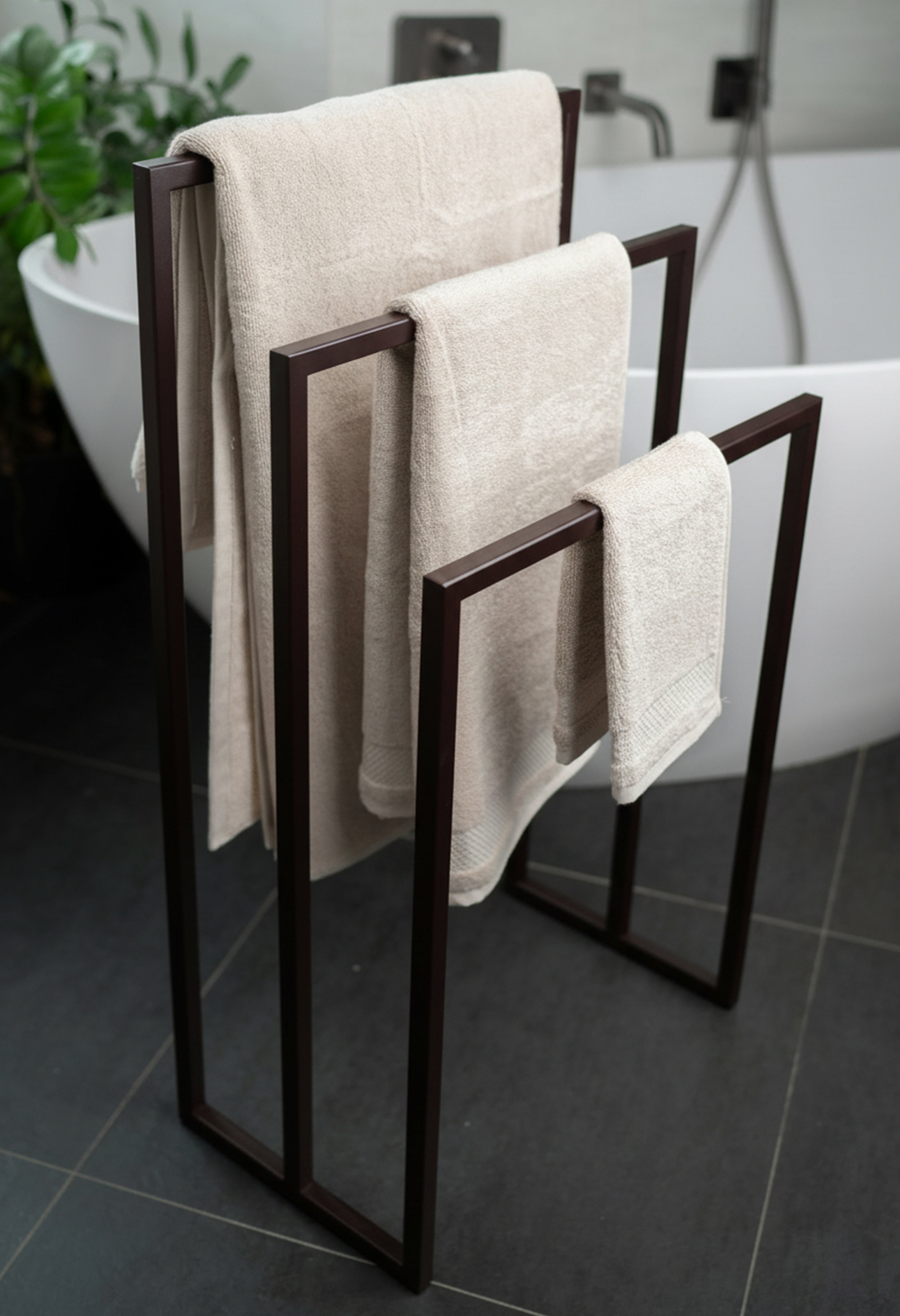 Freistehender Handtuchhalter in Marone braun aus Metall mit drei unterschiedlich hohen Stangen für Handtücher verschiedener Größen. Der Handtuchhalter steht vor einer modernen großen Badewanne in einem eleganten Badezimmer mit dunklen Fliesen