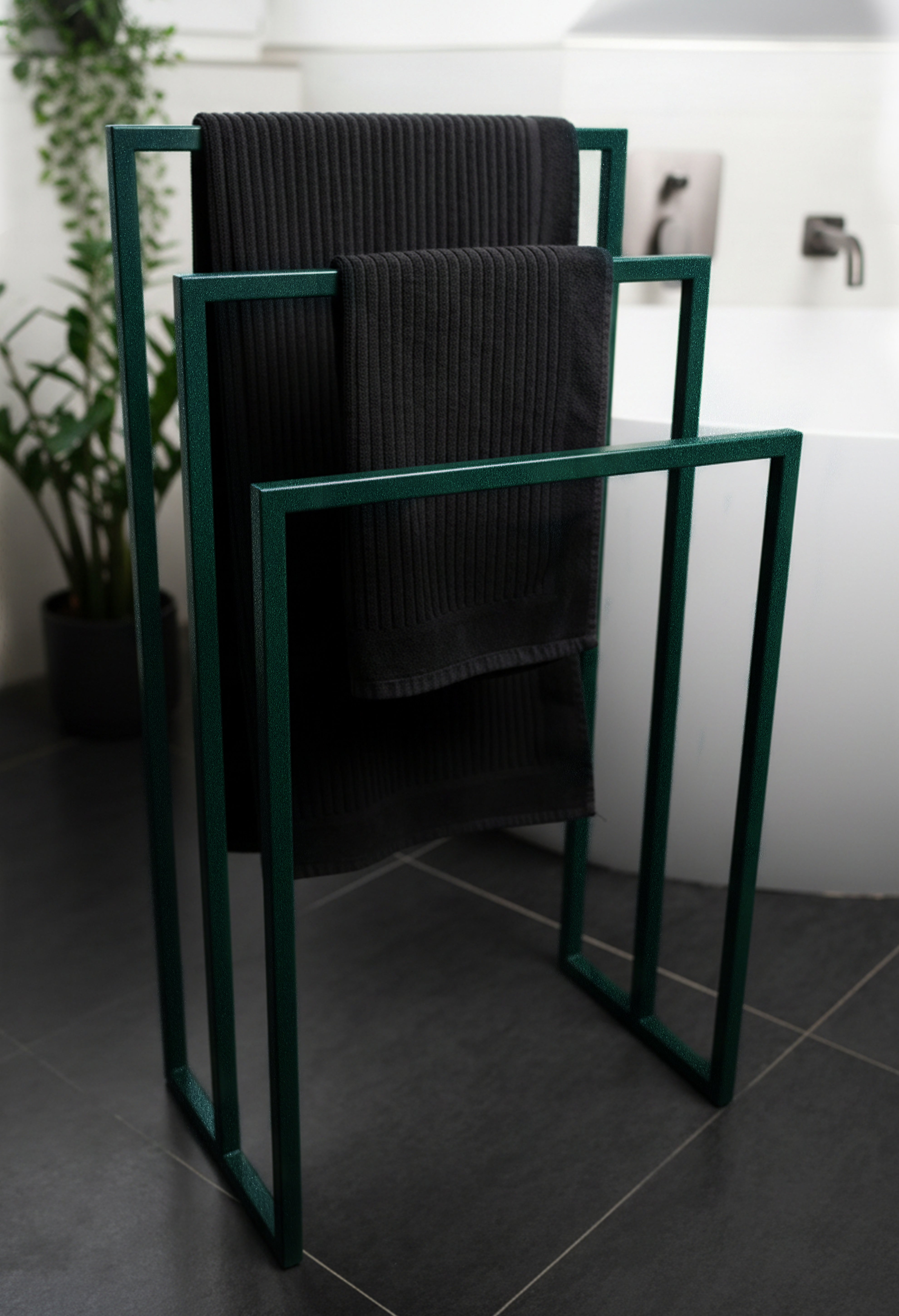 Freistehender Handtuchhalter in Sparkling Green aus Metall mit drei unterschiedlich hohen Stangen für Handtücher verschiedener Größen. Der Handtuchhalter steht vor einer modernen großen Badewanne in einem eleganten Badezimmer mit dunklen Fliesen