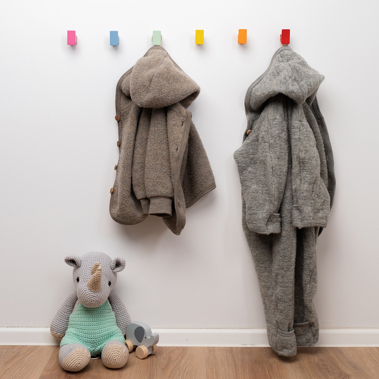 Bunte Haken auf niedirger Höhe an Wand angebracht für Kinder. An den Haken hängen zwei Kinderjäckchen und unter der Kindergarderobe sitzen ein kleines gehäkeltes Nashorn und ein Holz-Elefant in Pastellfarben.