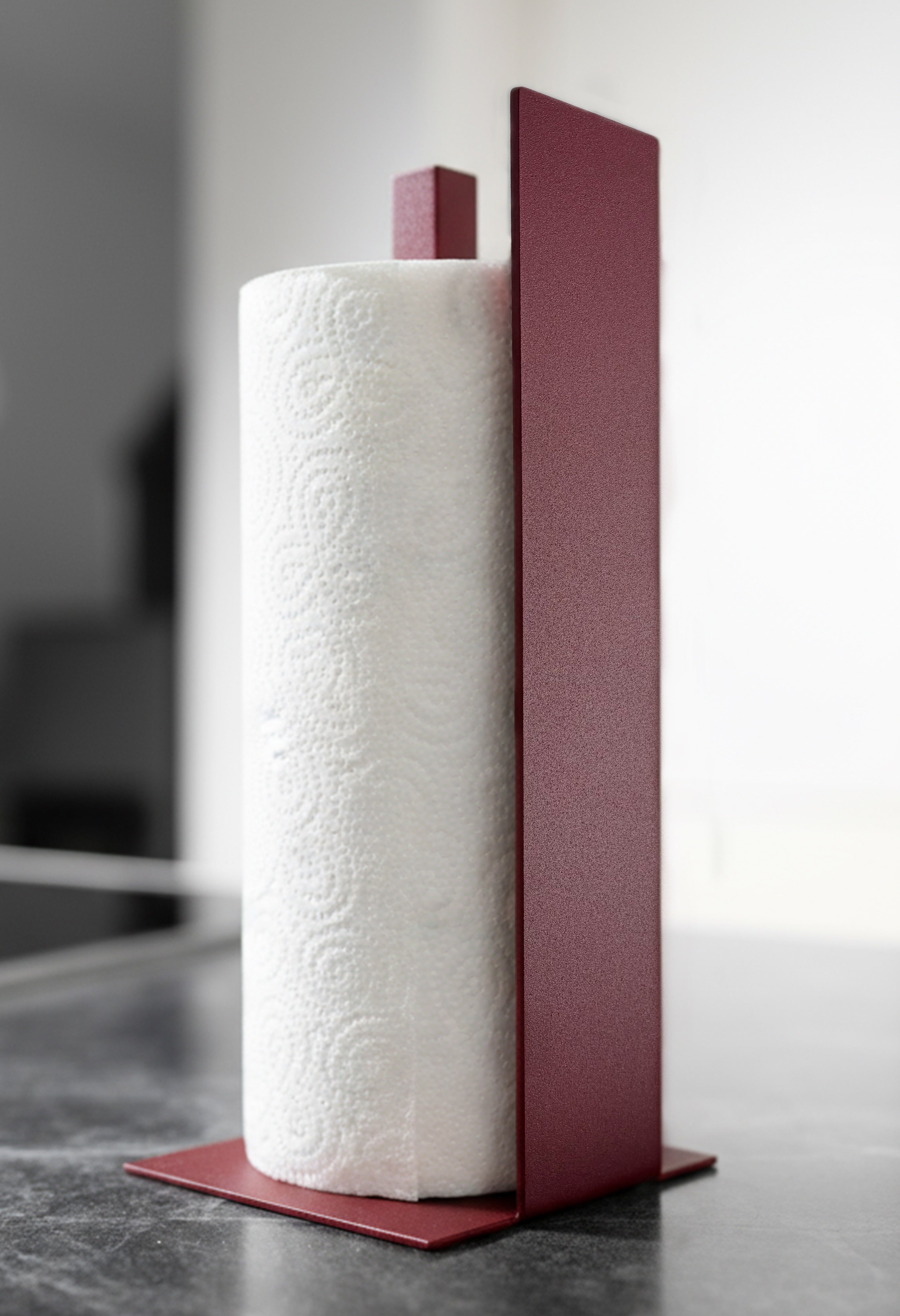 Küchenrollenhalter Granada pulverbeschichtet matt in Velvet Red, auf einer dunklen Marmorarbeitsfläche in einer hellen Küche. Schräge Ansicht von vorne.