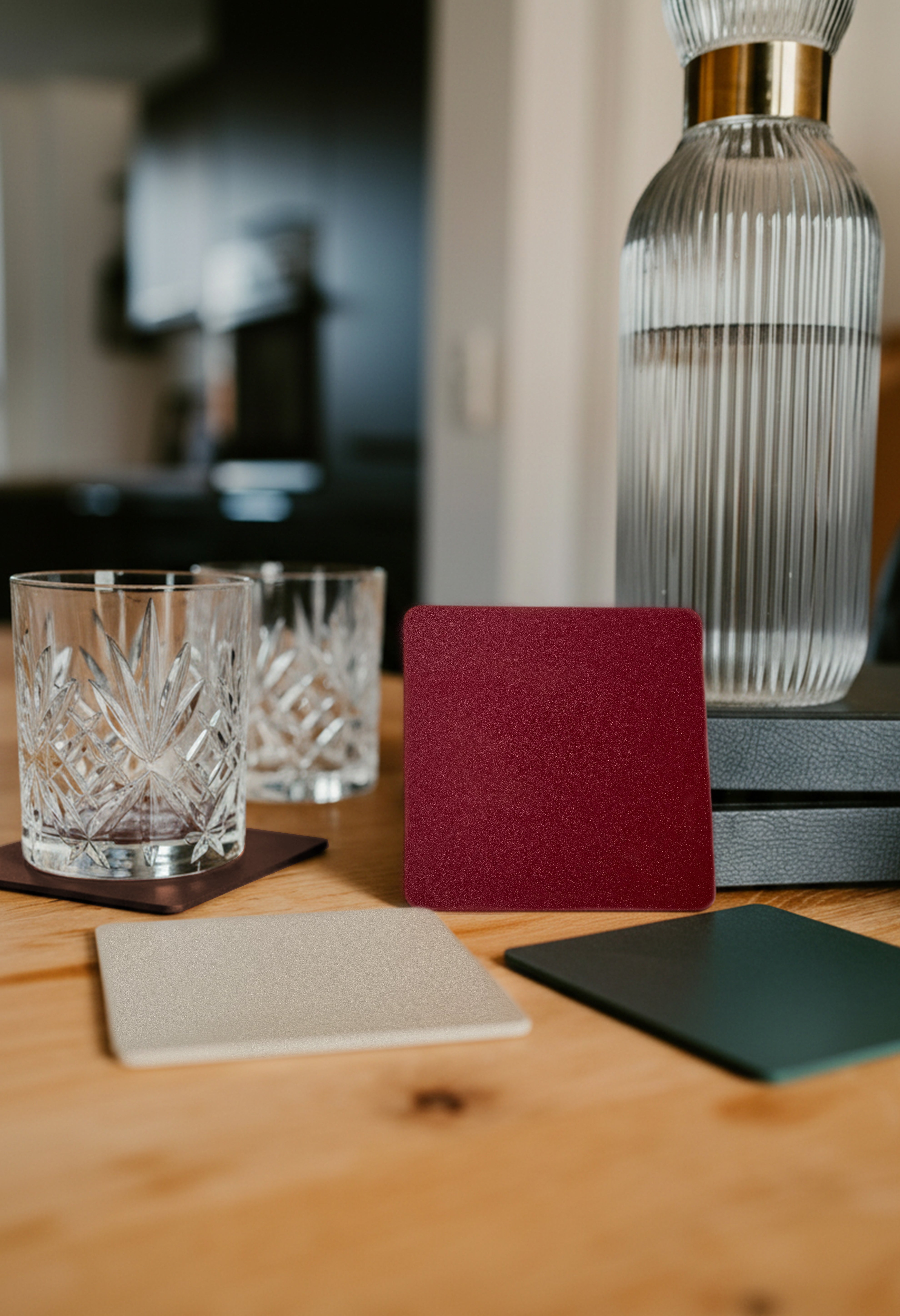 Velvet Red pulverbeschichteter Metall Untersetzer auf Holztisch lehnt an zwei Büchern auf welchem eine hochwertige Glaskaraffe steht. ZweiUntersetzer in champagner und sparkling green liegen davor. Im Hintergrund Kristallglas auf maronefarbenen Untersetzer.
