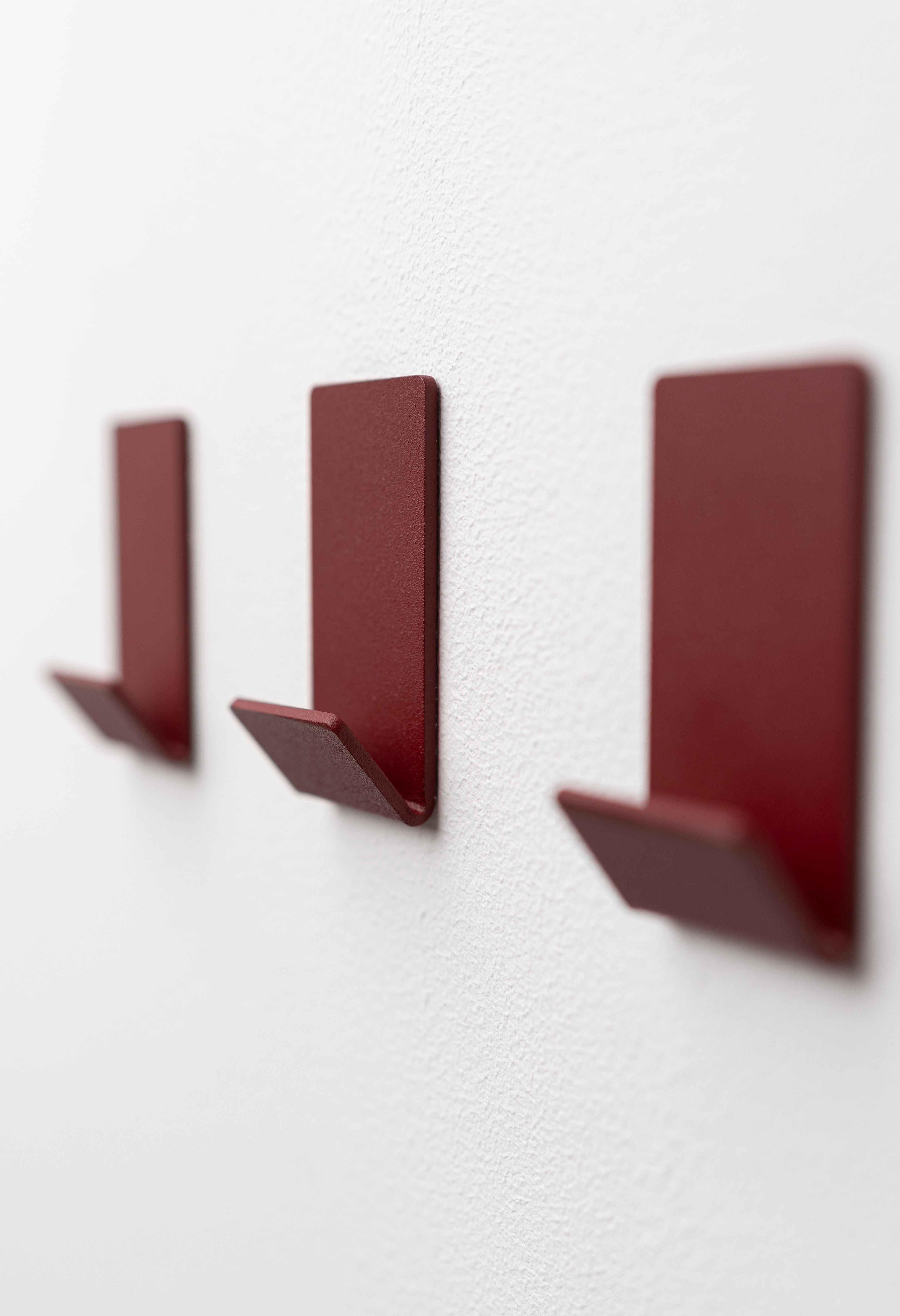 Wandhaken zum Kleben Ohne Bohren in Velvet Red pulverbeschichtet als dreier Set an weißer Wand.